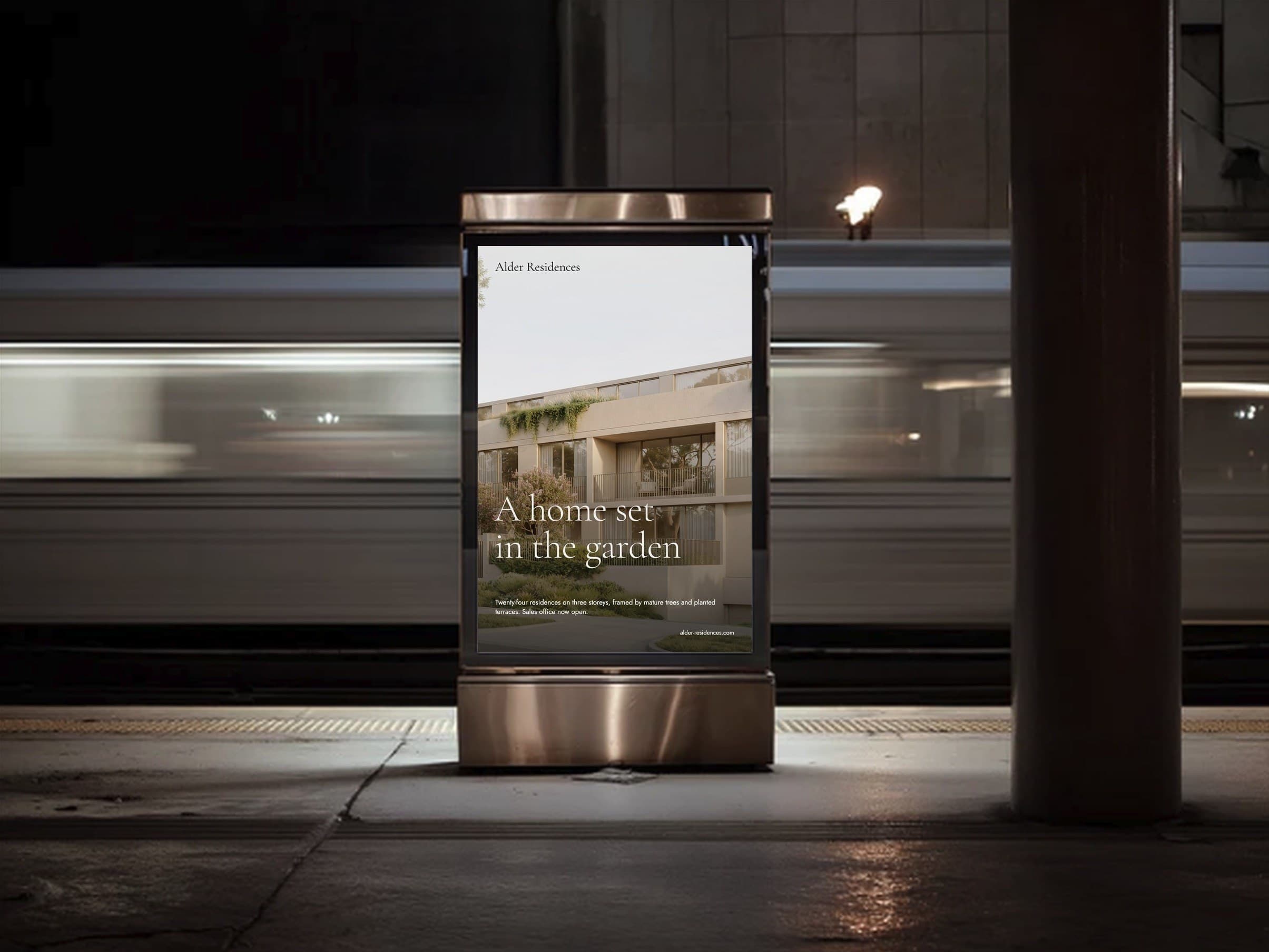 Alder Residences poster in a lit display case on a station platform, a train blurred behind it